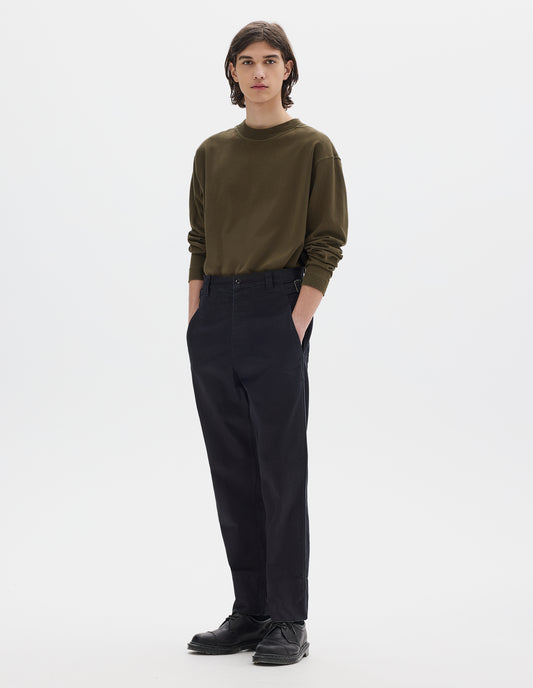 Full body image, front view of model wearing dark blue trousers with belt loops and side cinches, slant pockets, zip fly and corozo button fastening, deep stitch hem. Styled with brown long sleeve t-shirt tucked in and black leather shoes.
