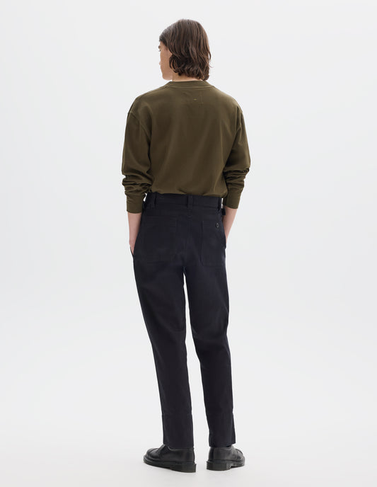 Full body image, back view of model wearing dark blue trousers with belt loops and side cinches, corozo button-through patch back pockets. Styled with brown long sleeve t-shirt tucked in and black leather shoes.