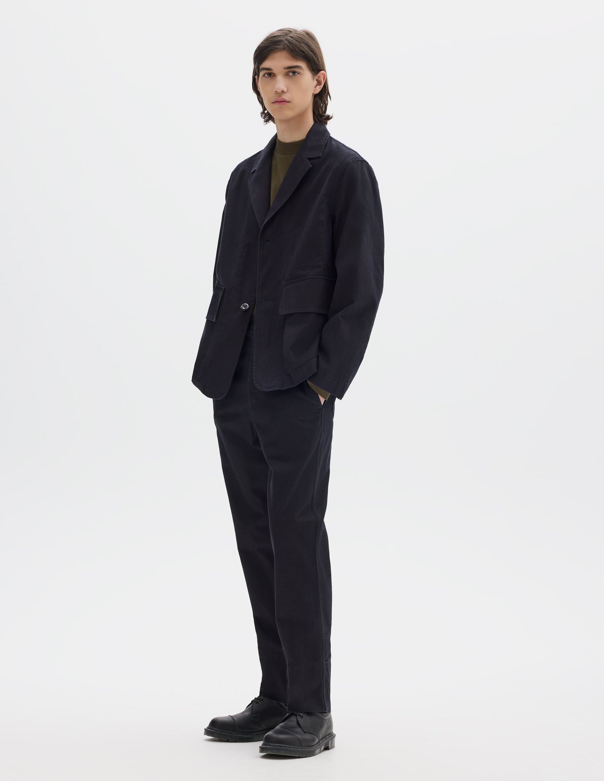 Full body image, front view of model wearing indigo blue twill blazer with corozo button fastening lapel, stitch through chest pocket and flap patch pockets. Styled over brownish green jersey and with matching indigo blue twill tapered trousers and black leather shoes.