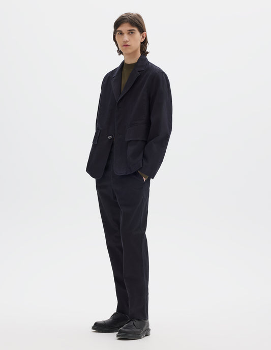 Full body image, front view of model wearing indigo blue twill blazer with corozo button fastening lapel, stitch through chest pocket and flap patch pockets. Styled over brownish green jersey and with matching indigo blue twill tapered trousers and black leather shoes.