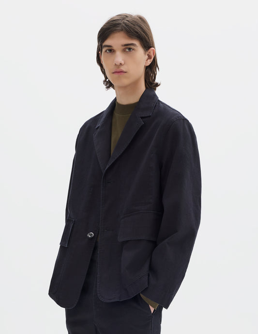 Cropped body image, front view of model wearing indigo blue twill blazer with corozo button fastening lapel, stitch through chest pocket and flap patch pockets. Styled over brownish green jersey and with matching indigo blue twill tapered trousers.