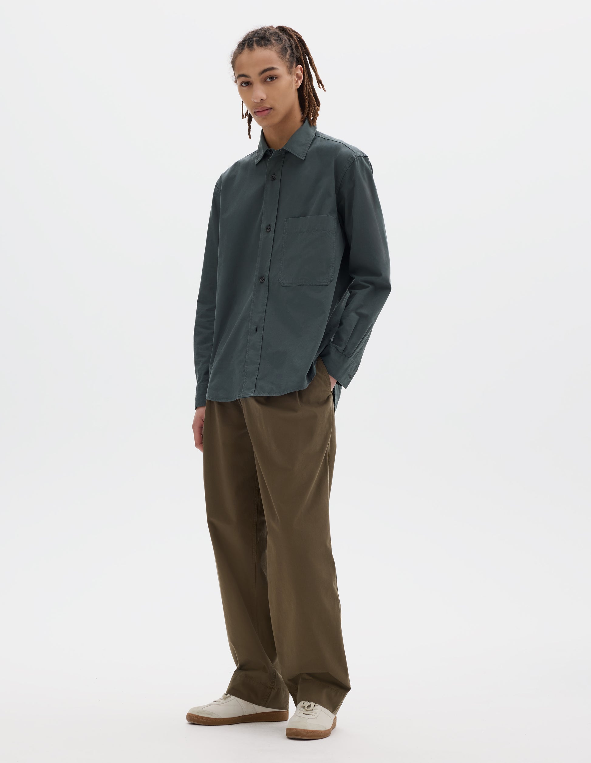 Full body image, front view of model wearing relaxed cut dark grey shirt with raised placket, large chest pocket, side gusset stitch detailing. Styled with khaki pleated trousers and white suede trainers.