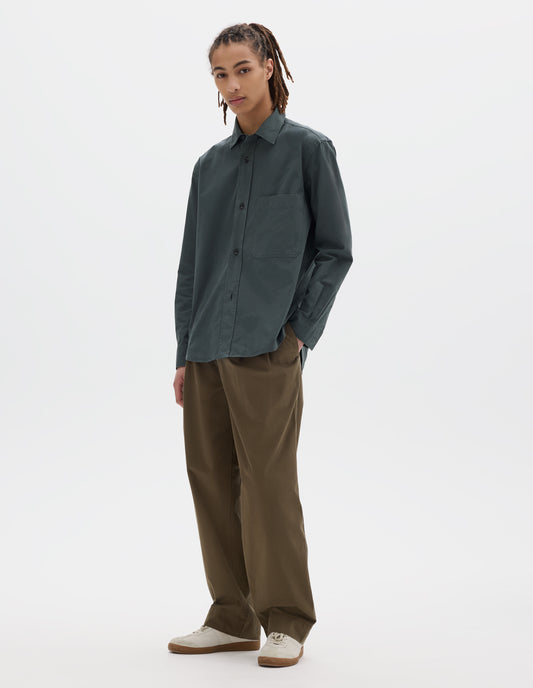 Full body image, front view of model wearing relaxed cut dark grey shirt with raised placket, large chest pocket, side gusset stitch detailing. Styled with khaki pleated trousers and white suede trainers.