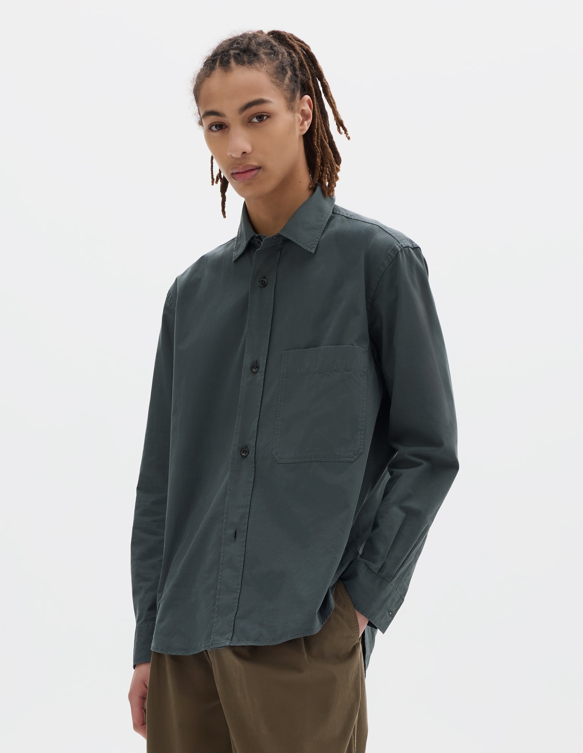 Cropped image, front view of model wearing relaxed cut dark grey shirt with raised placket, large chest pocket, side gusset stitch detailing. Styled with khaki pleated trousers.