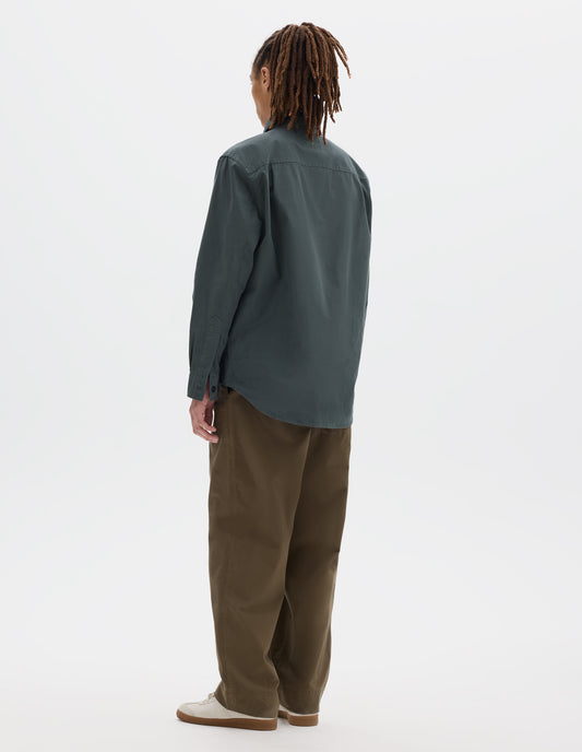 Full body image, back view of model wearing relaxed cut dark grey shirt with side gusset stitch detailing and pleats on the back yoke. Styled with khaki trousers and white suede trainers.