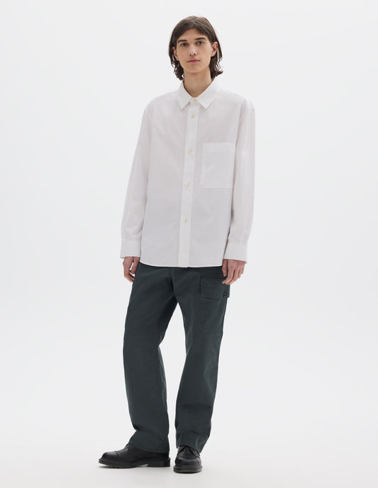 Full body image, front view of model wearing relaxed white shirt with raised placket, chest pocket and corozo brace button fastening. Styled with dark greenish cargo trousers and black leather shoes.