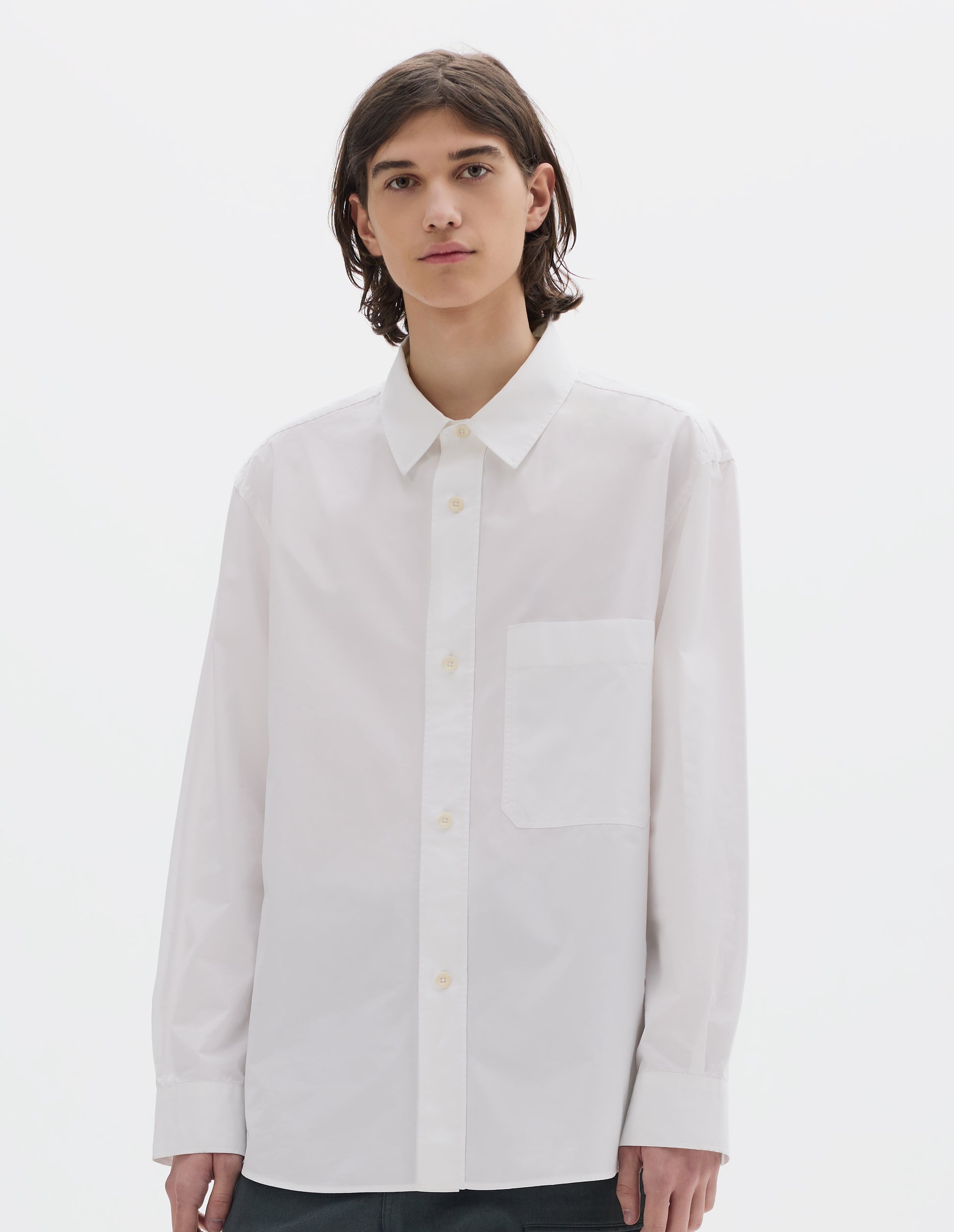 Cropped body image, front view of model wearing relaxed white shirt with  raised placket, chest pocket and corozo brace button fastening. Styled with dark greenish trousers.