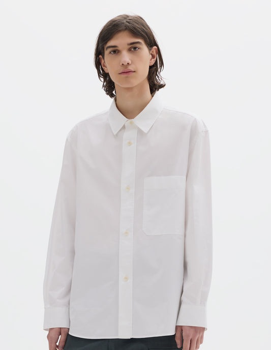 Cropped body image, front view of model wearing relaxed white shirt with  raised placket, chest pocket and corozo brace button fastening. Styled with dark greenish trousers.
