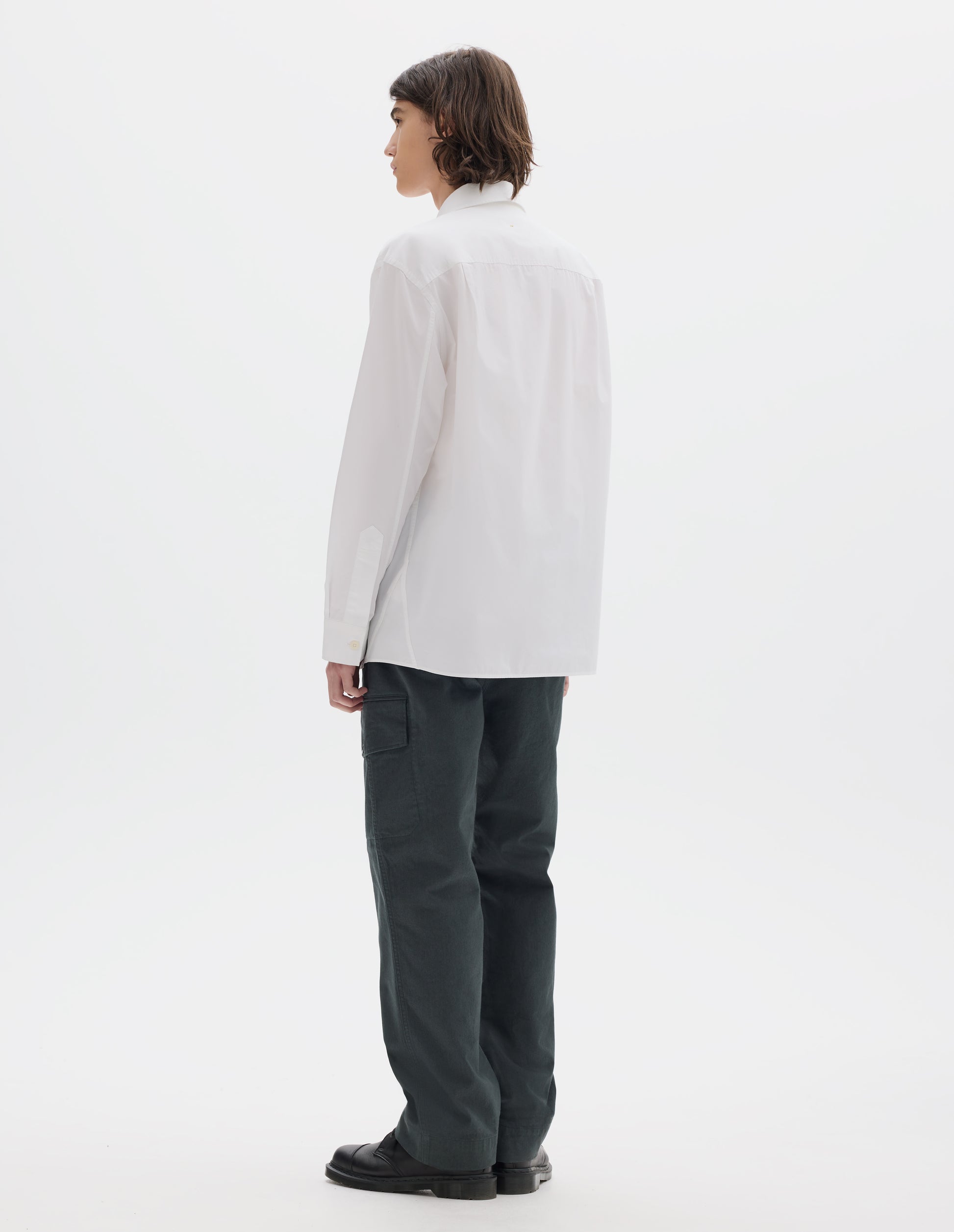 Full body image, back view of model wearing relaxed white shirt with pleats on back yoke and corozo brace buttons on cuffs. Styled with dark greenish cargo trousers and black leather shoes.