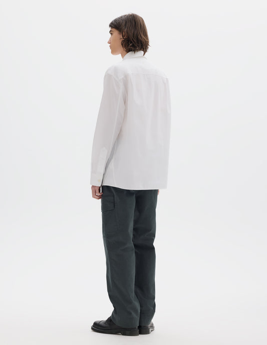 Full body image, back view of model wearing relaxed white shirt with pleats on back yoke and corozo brace buttons on cuffs. Styled with dark greenish cargo trousers and black leather shoes.