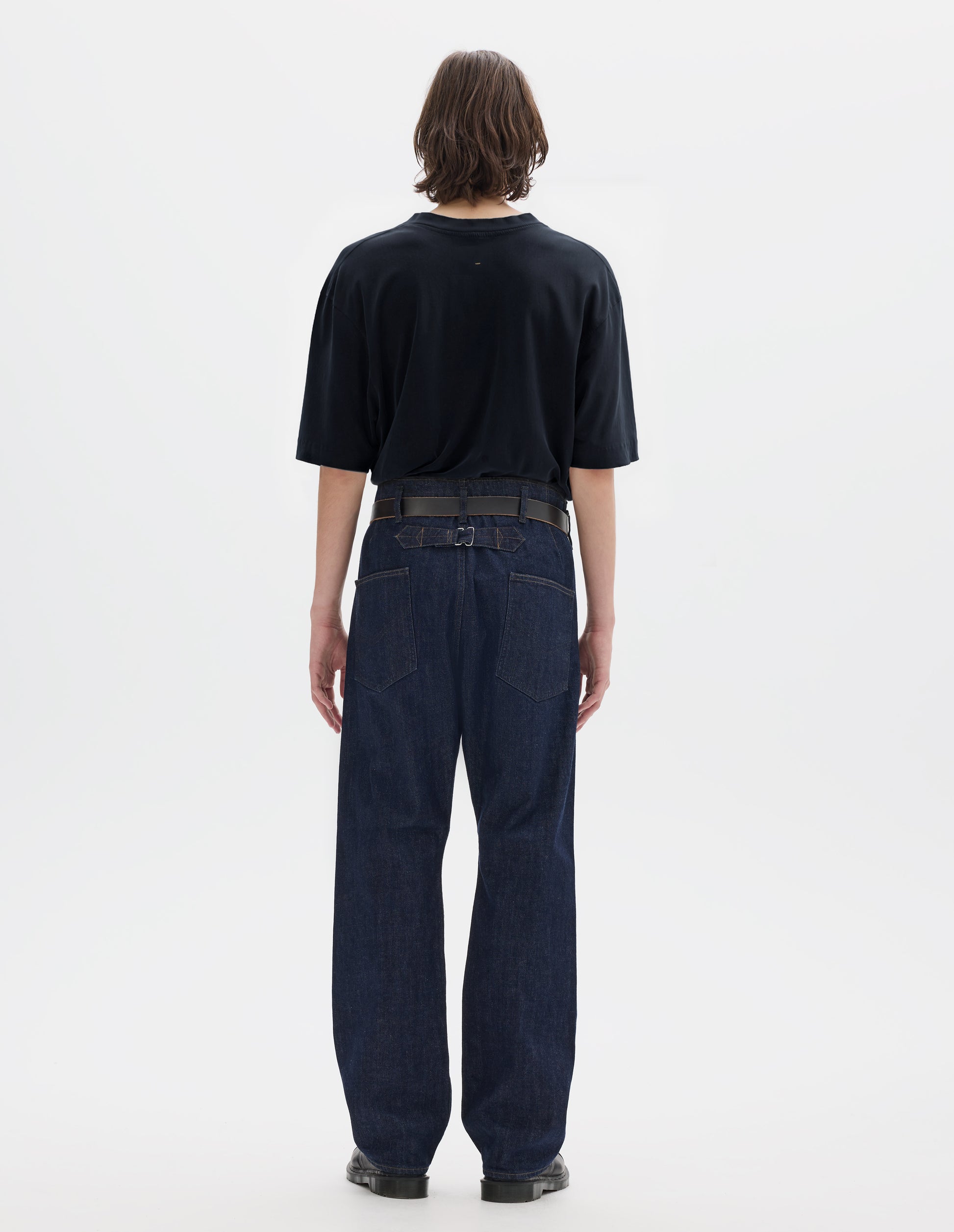 Full body image, back view of model wearing navy blue short sleeve cotton linen t-shirt with contrast rib on neck, worn tucked in to blue denim trousers with black leather belt and black leather shoes.
