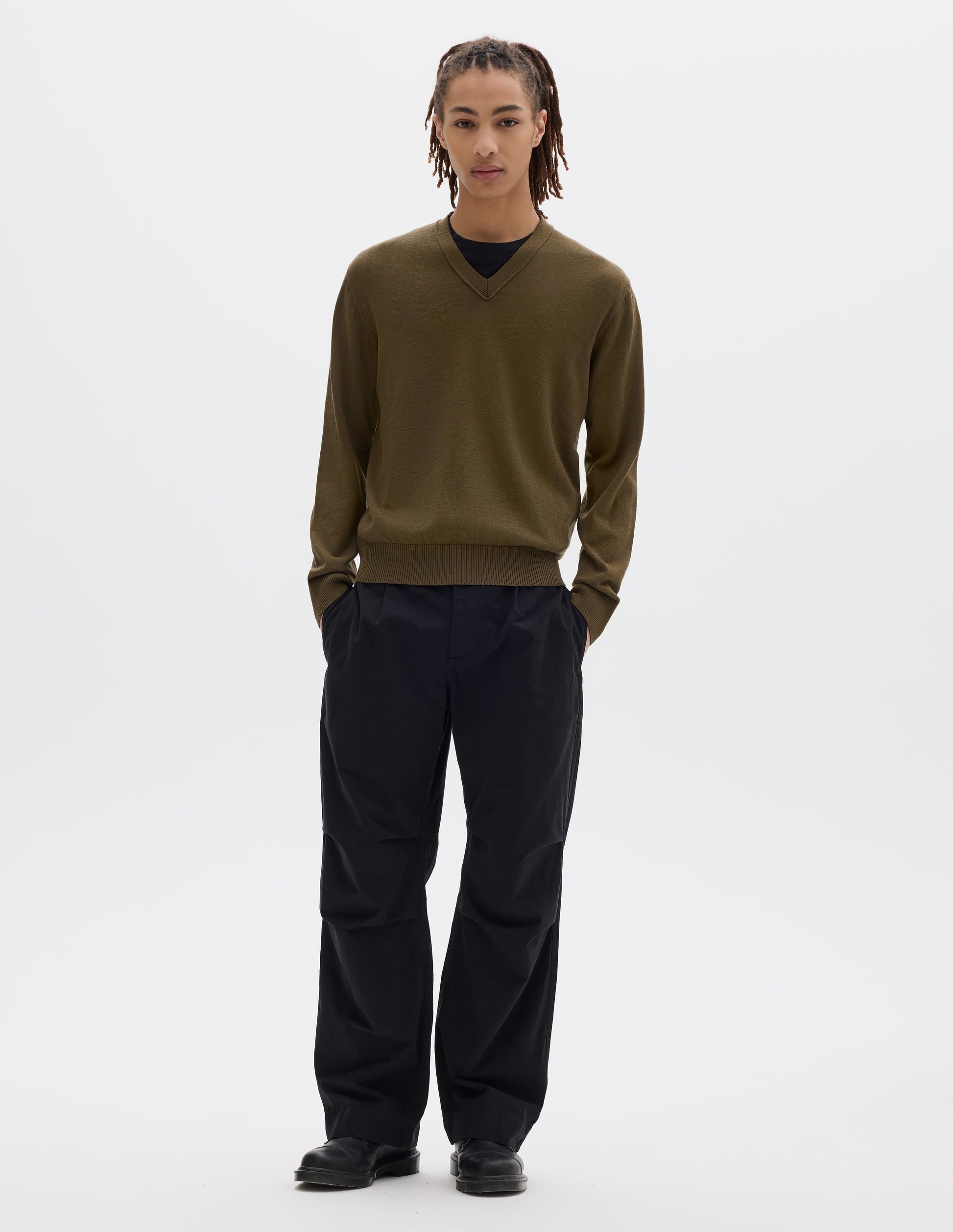 Full body image, front view of model wearing green brown fine wool jumper, with a v neck, flatlock detailing on the sleeves and rib cuffs and hem. Styled with a black t shirt, black pleated trousers and black leather shoes.