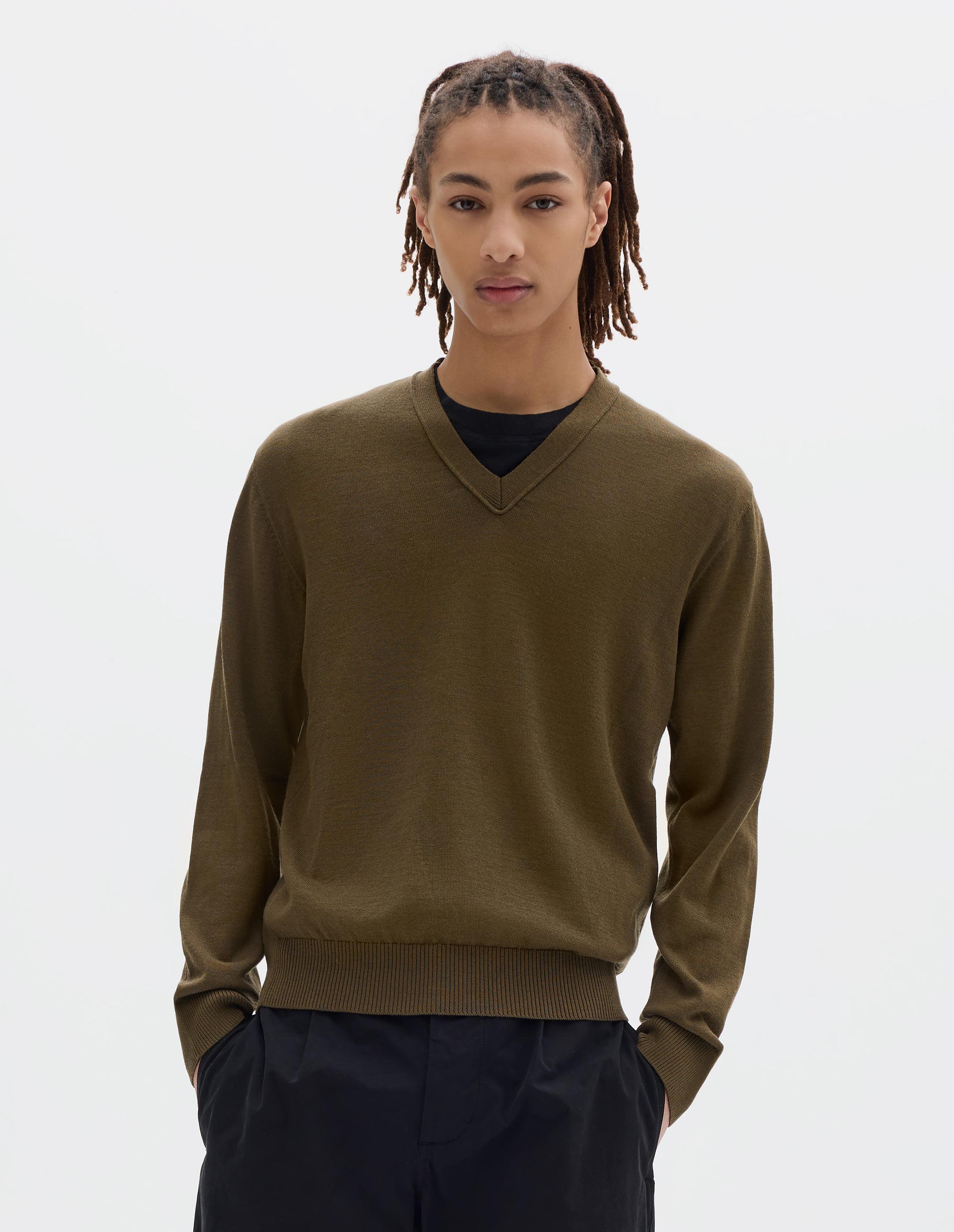 Cropped body image, front view of model wearing green brown fine wool jumper with a v neck, finished with rib cuffs and hem. Styled over a black t shirt and black trousers. 