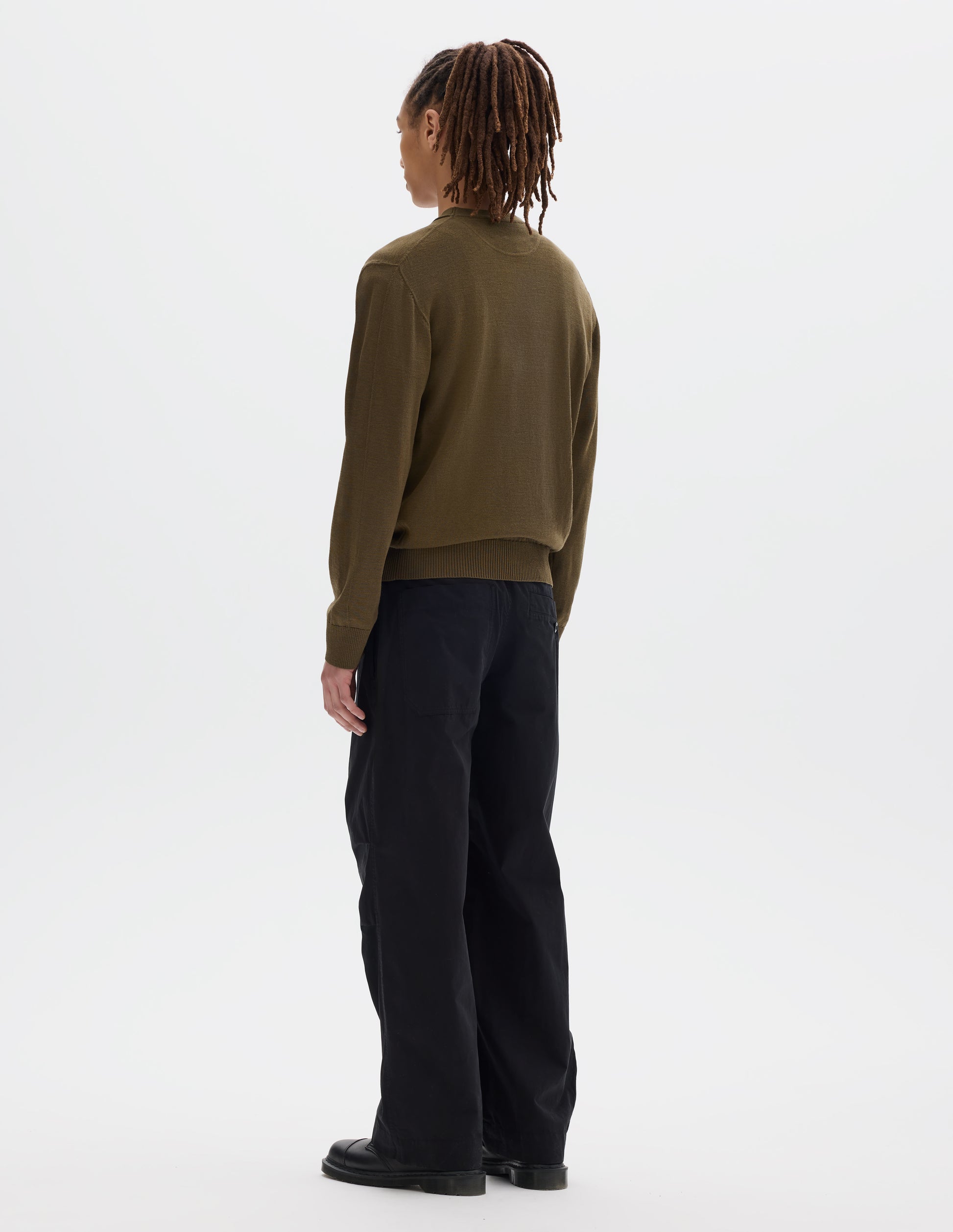 Full body image, back view of model wearing green brown fine wool jumper, with a half-moon yoke at the back neck, flatlock detailing on the sleeves and rib on cuffs and hem. Styled with a black t shirt, black trousers and black leather shoes.