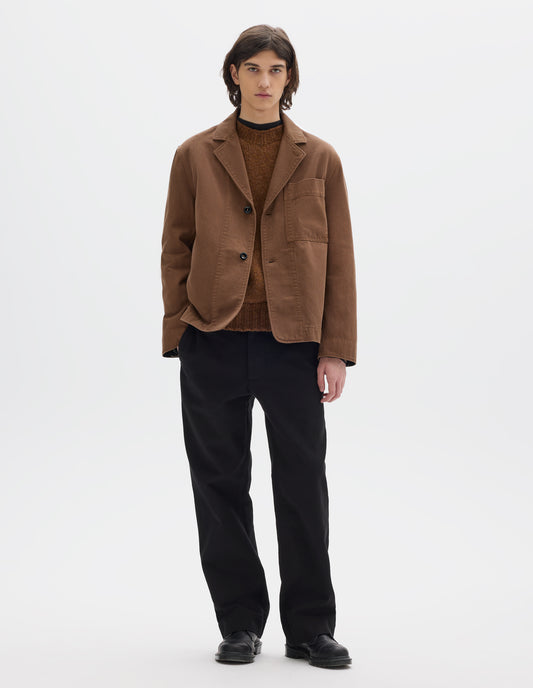 Full body image, front view of model wearing a brown cotton blazer, constructed with a button fastening lapel, large patch chest pocket and stitch-through facing. Styled over a brown jumper and black jersey. Worn with black cotton trousers and black leather shoes.