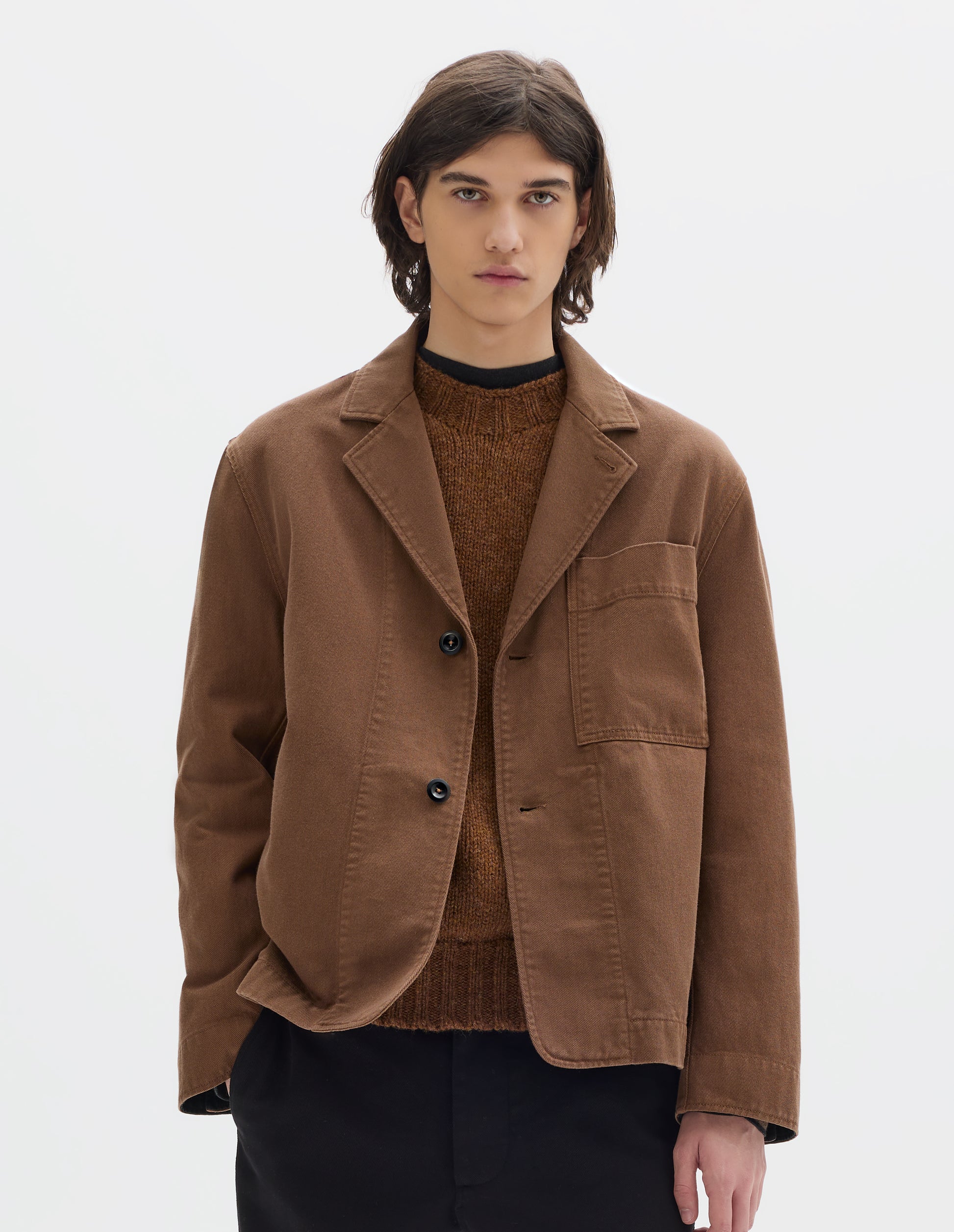 Cropped body image, front view of model wearing a brown cotton blazer, constructed with a button fastening lapel, large patch chest pocket and stitch-through facing. Styled over a brown jumper and black jersey. Worn with black cotton trousers.