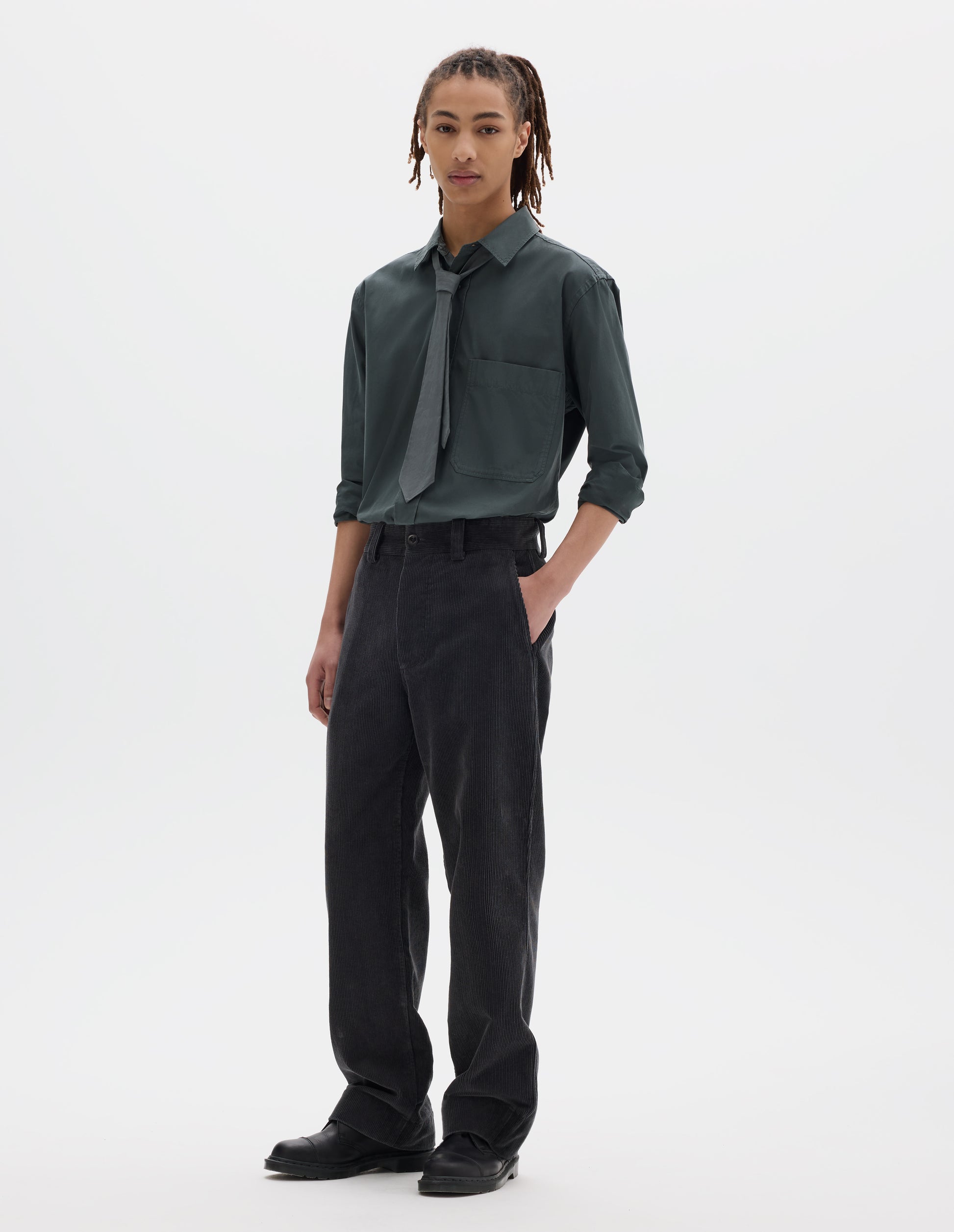 Full body image, front view of model wearing dark grey cotton corduroy trousers with belt loops, a button fastening and slanted side entry pockets. Styled with greenish grey shirt tucked in and matching tie, with black leather shoes.