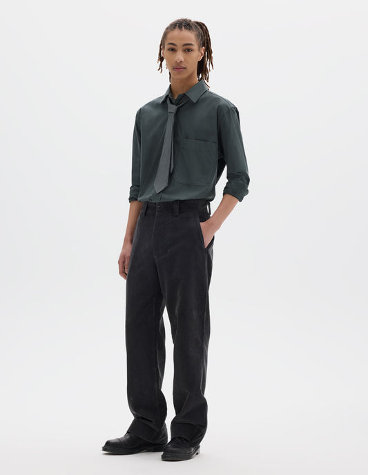 Full body image, front view of model wearing dark grey cotton corduroy trousers with belt loops, a button fastening and slanted side entry pockets. Styled with greenish grey shirt tucked in and matching tie, with black leather shoes.