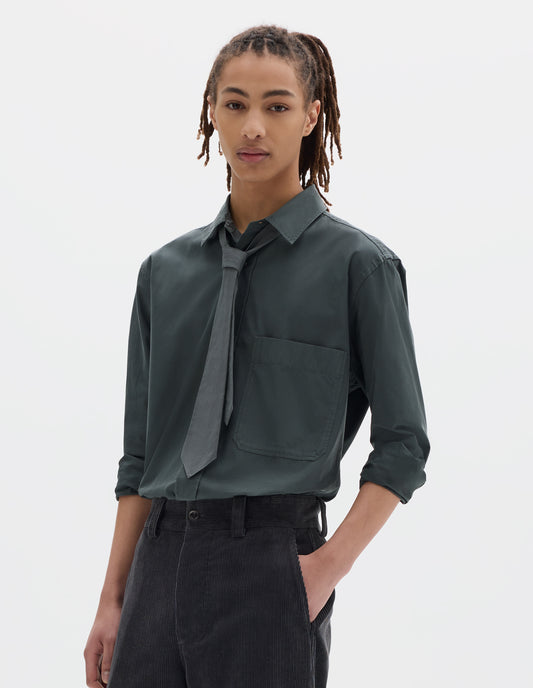 Cropped body image, front view of model wearing a blue green shirt with a front patch pocket, sleeves rolled up, styled with a greyish green tie and a dark greyish flat front cotton trousers.