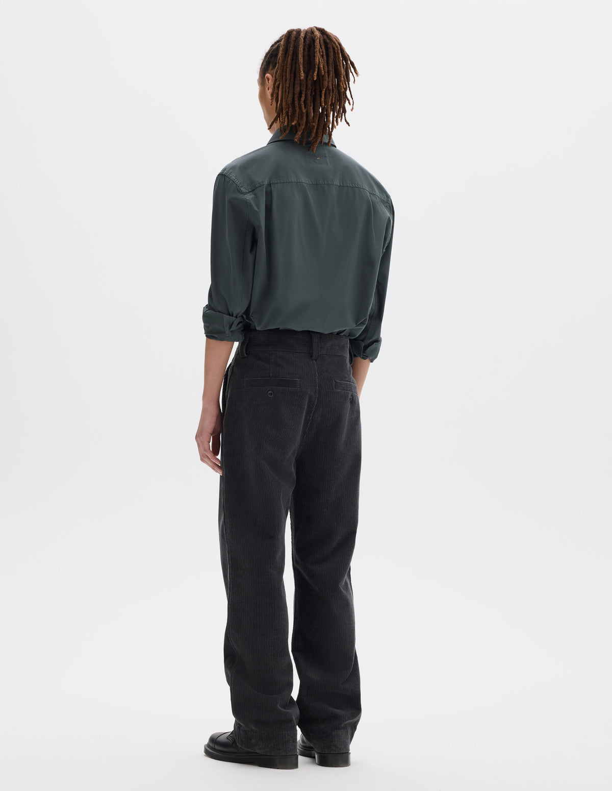 Full body image, back view of model wearing dark grey cotton corduroy trousers with belt loops and two welt back button fastening pockets. Styled with greenish grey shirt tucked in and black leather shoes.