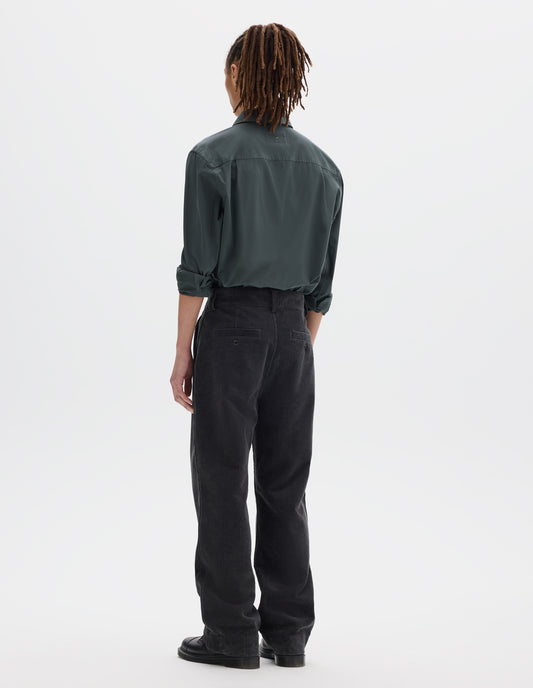 Full body image, back view of model wearing dark grey cotton corduroy trousers with belt loops and two welt back button fastening pockets. Styled with greenish grey shirt tucked in and black leather shoes.