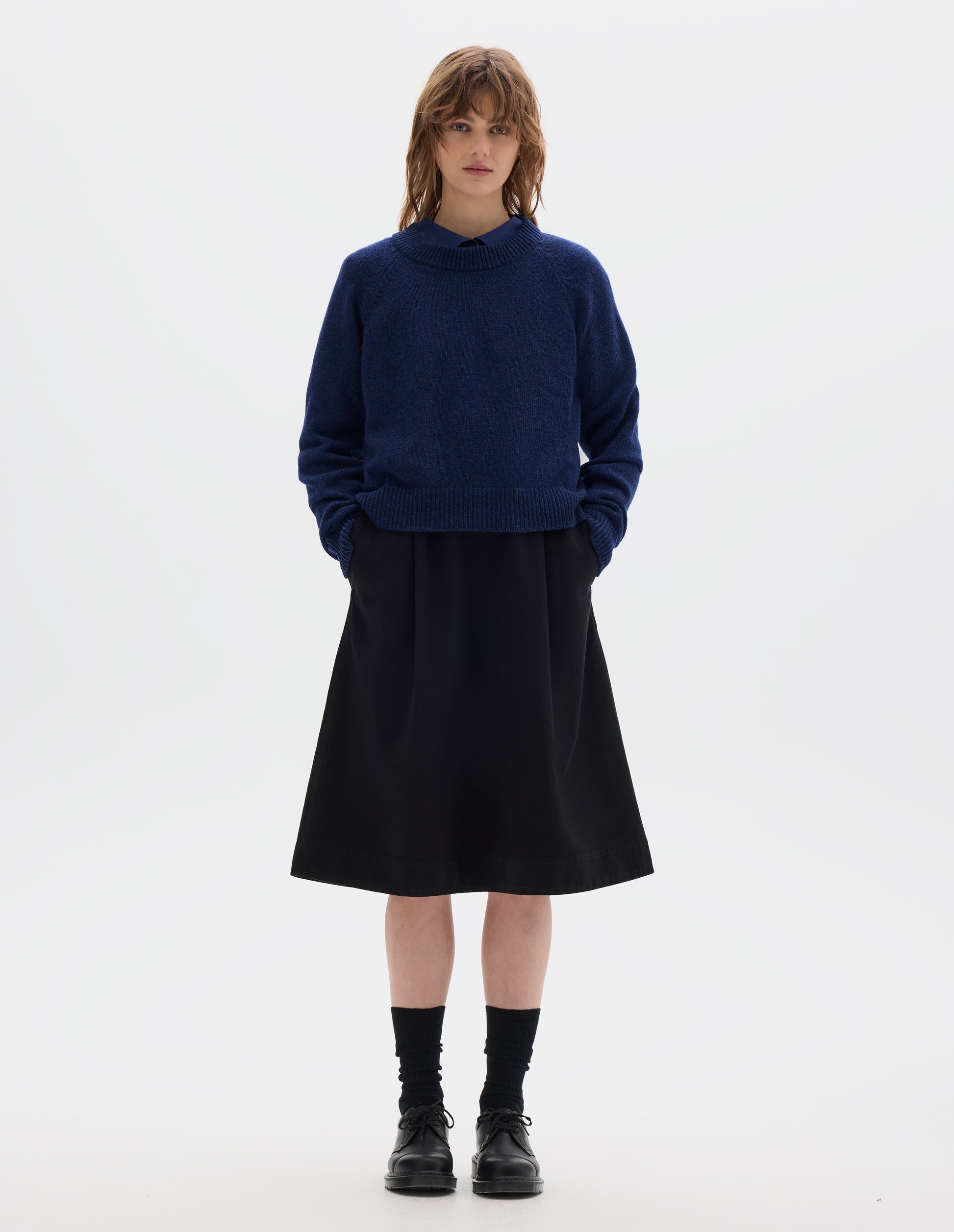 Full body image, front view of model wearing blue lambswool jumper, with crew neck, raglan sleeves, and ribbed neck, cuffs and hem. Styled with blue shirt, black skirt, black socks and black shoes.
