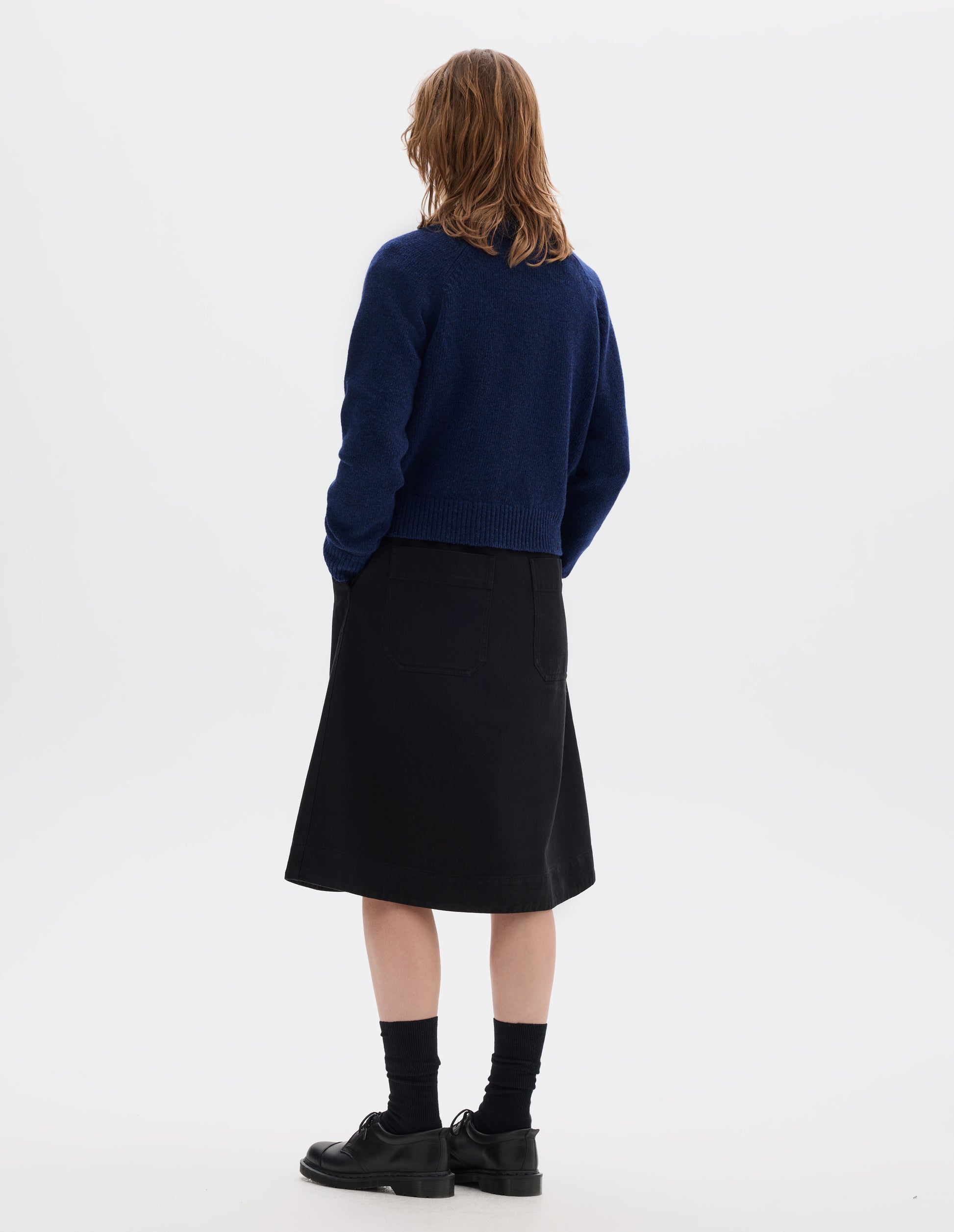 Full body image, back view of model wearing blue lambswool jumper, with crew neck, raglan sleeves, and ribbed neck, cuffs and hem. Styled with black skirt, black socks and shoes.
