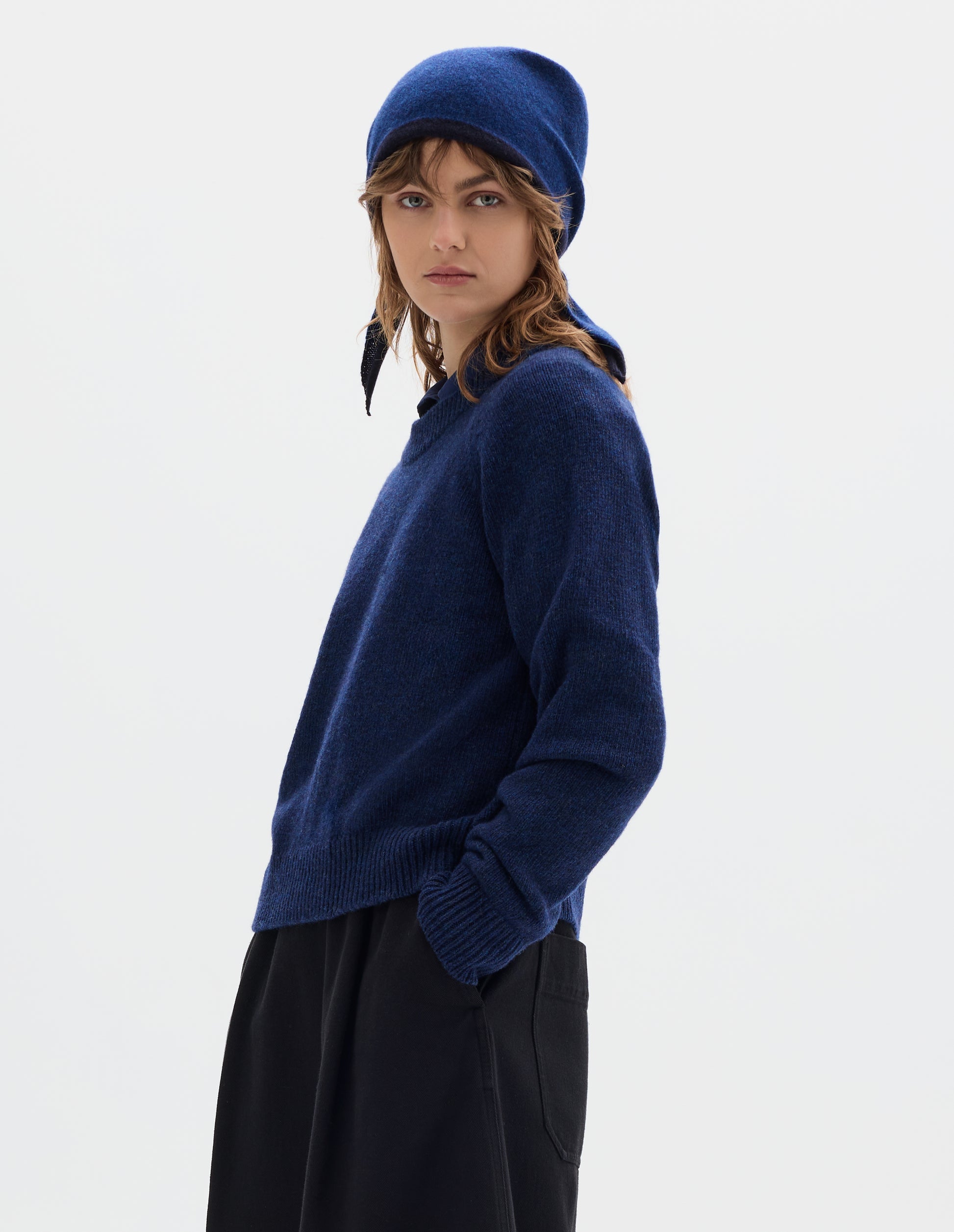 Cropped body image, front view of model wearing a blue wool triangle scarf with navy edge, tied on model's head. Styled with blue crew neck jumper and black pleated trousers.