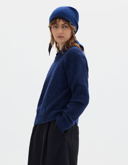 Cropped body image, front view of model wearing a blue wool triangle scarf with navy edge, tied on model's head. Styled with blue crew neck jumper and black pleated trousers.