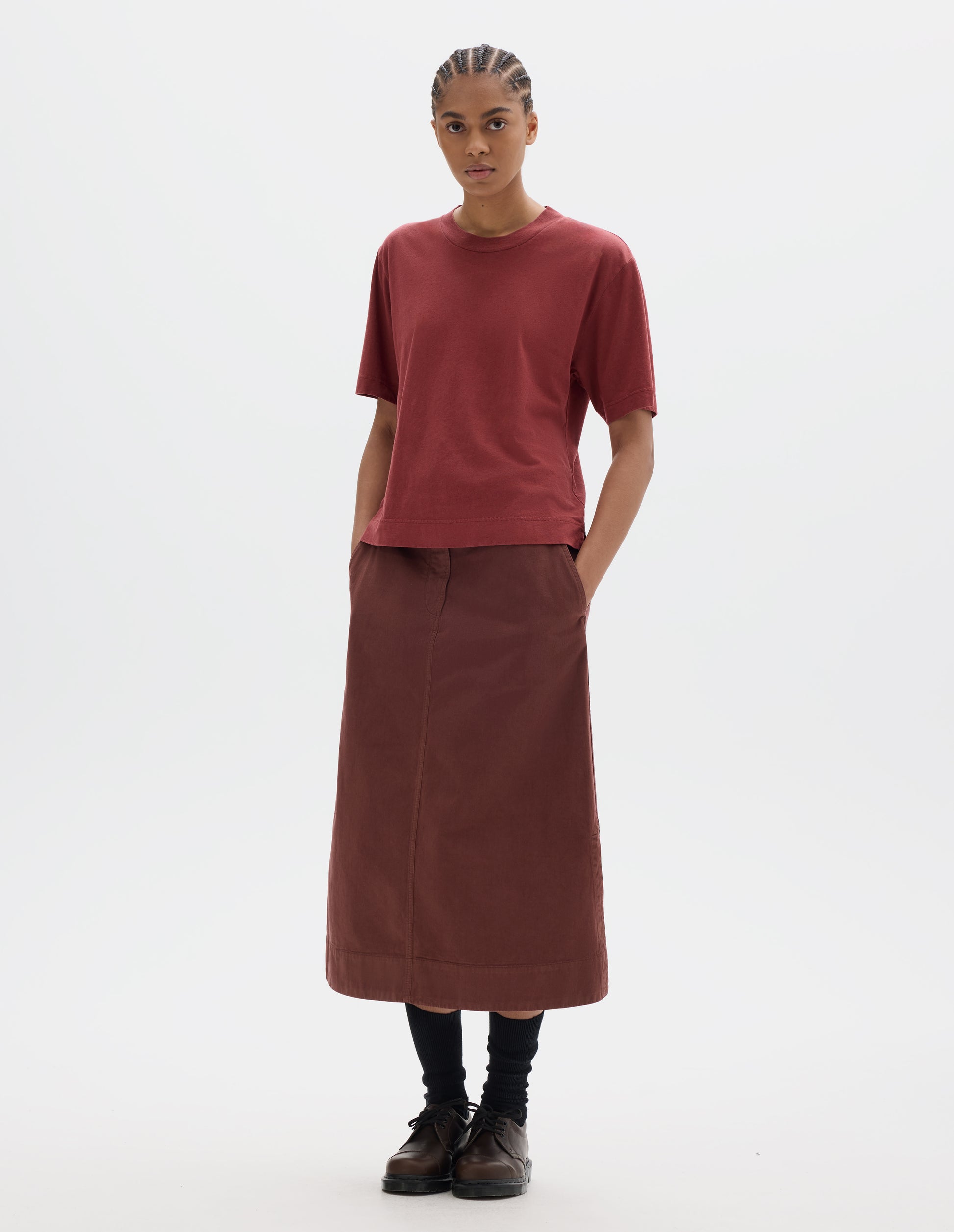 Full body image, front view of model wearing a boxy red short sleeve linen cotton t shirt with vent split hem. Styled with a midi red-brown cotton skirt, black socks and brown leather shoes.