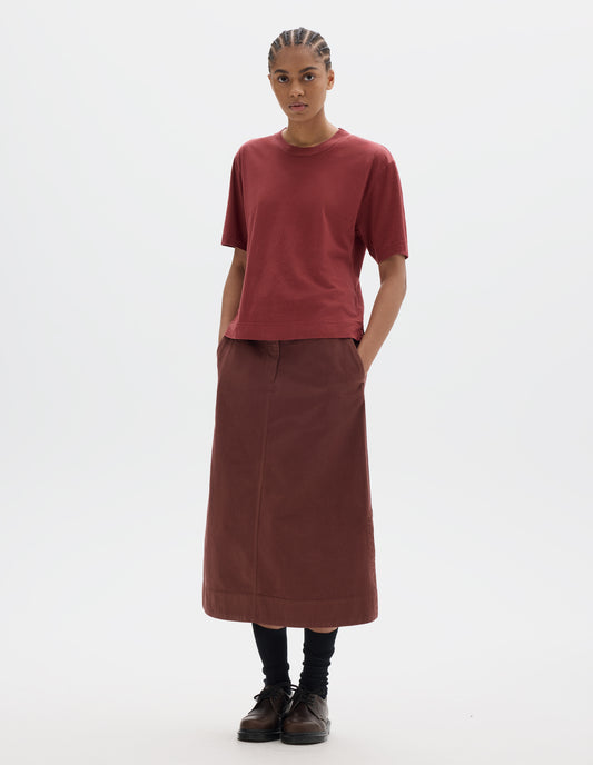 Full body image, front view of model wearing a boxy red short sleeve linen cotton t shirt with vent split hem. Styled with a midi red-brown cotton skirt, black socks and brown leather shoes.