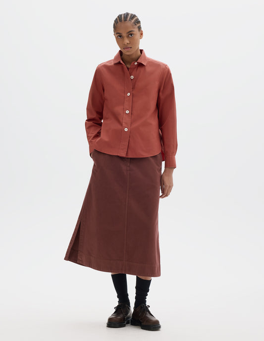 Full body image, front view of model wearing a red cotton poplin shirt with light corozo buttons, stitch-through raised placket, long sleeves with a button-through cuffs and a curved hem. Styled with a midi red-brown skirt, black socks and brown leather shoes.