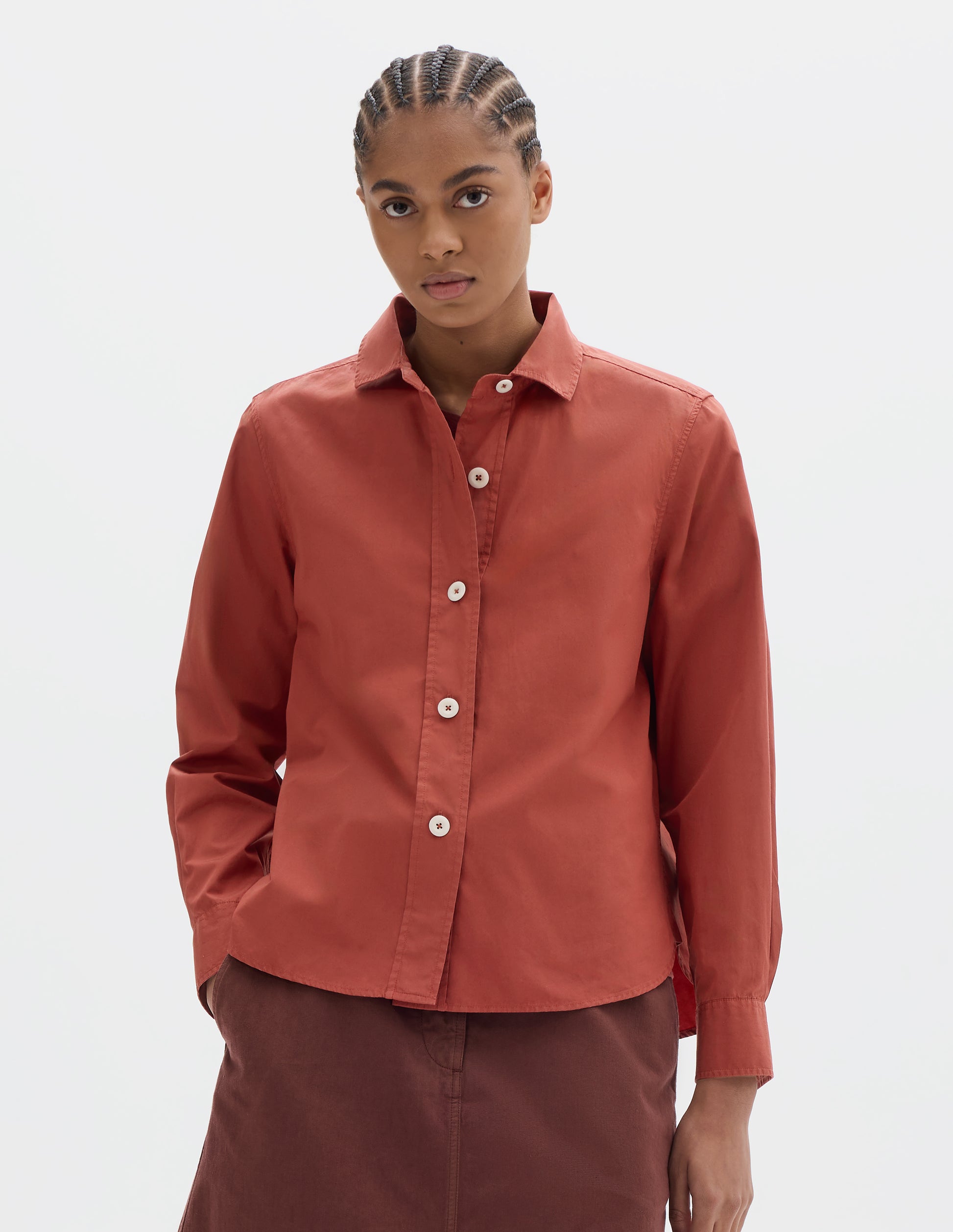 Cropped body image, front view of model wearing a red cotton poplin shirt with light corozo buttons, stitch-through raised placket, long sleeves with a button-through cuffs and a curved hem. Styled with a red-brown skirt.