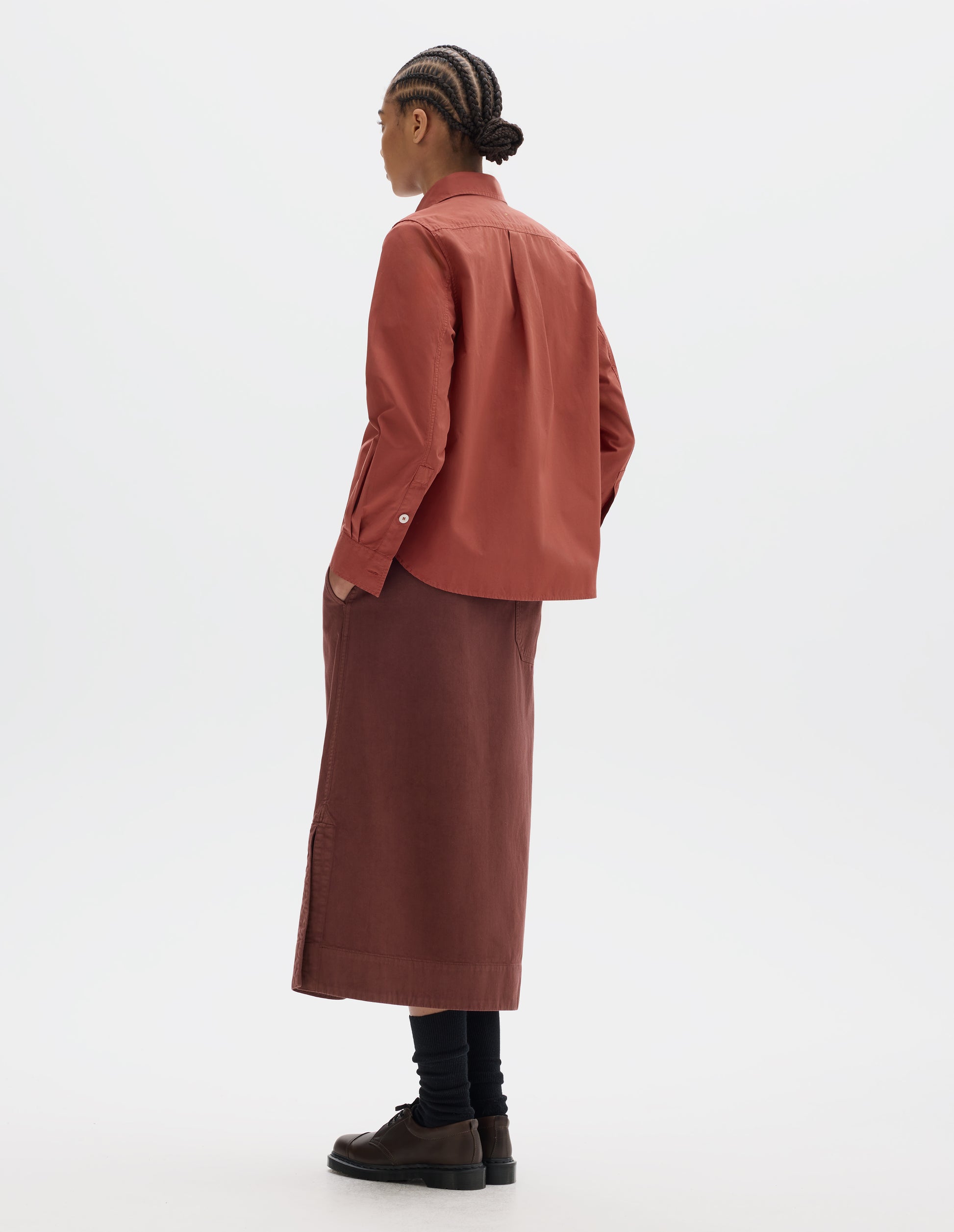 Full body image, back view of model wearing a red cotton poplin shirt with long sleeves with a button-through cuffs, a box pleat on the back yoke and a curved hem. Styled with a red-brown midi skirt, black socks and brown leather shoes.