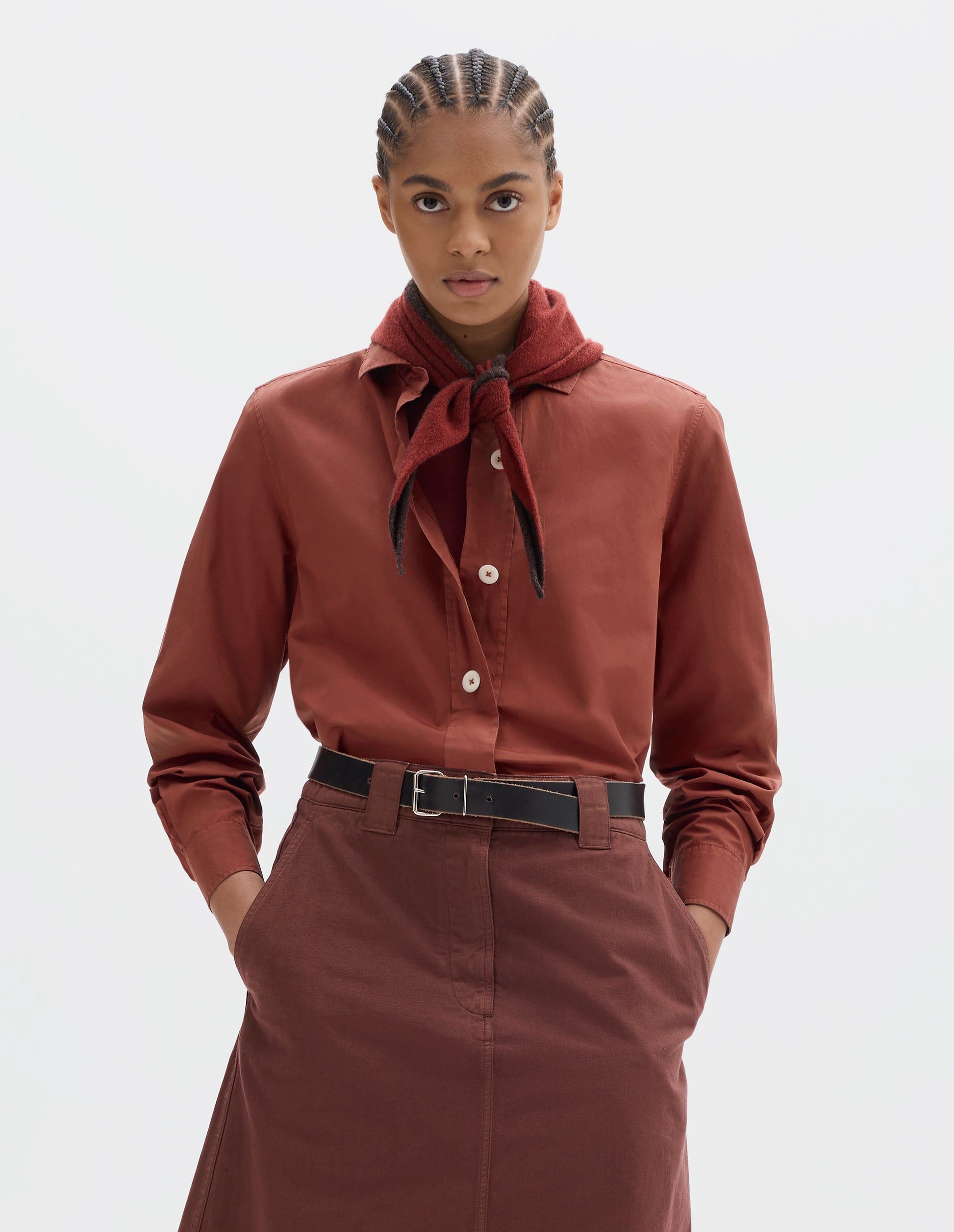 Cropped body image, front view of model wearing a orangey red wool triangle scarf eith dark brown edge, tied around her neck, worn with a red cotton shirt with light buttons and styled with a reddish brown skirt and black leather belt.