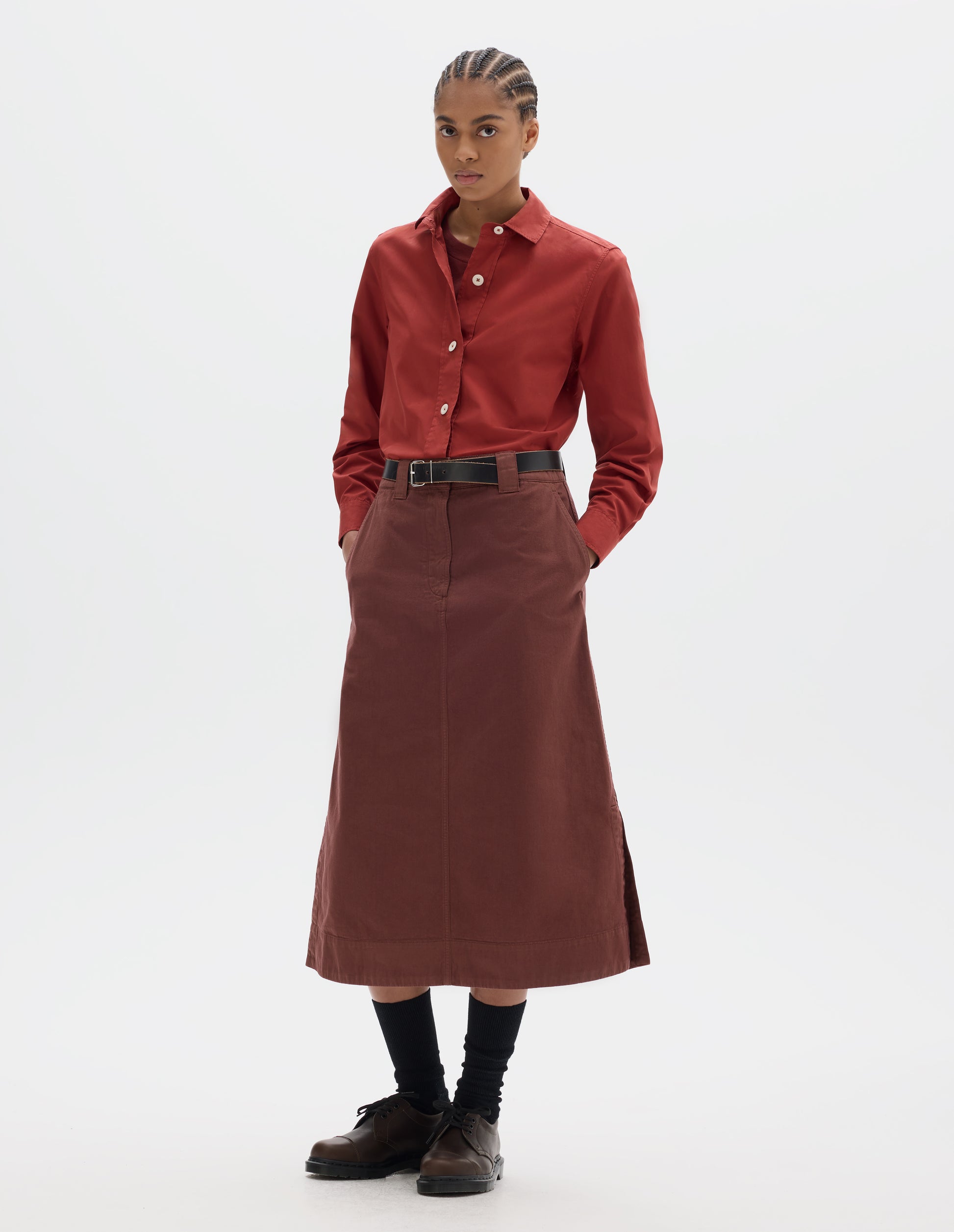 Full body image, front view of model wearing red A line skirt with side splits, double hook and bar fastening waistband, belt loops and slant pockets. Styled with red t shirt and red shirt tucked in, black leather belt and black socks with brown leather shoes. 