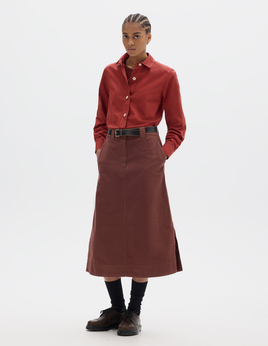 Full body image, front view of model wearing red A line skirt with side splits, double hook and bar fastening waistband, belt loops and slant pockets. Styled with red t shirt and red shirt tucked in, black leather belt and black socks with brown leather shoes. 