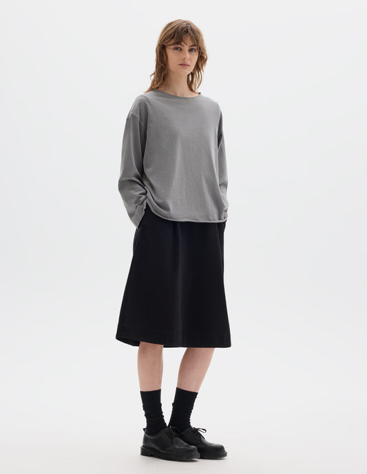 Full body image, front view of model wearing a wide neckline grey long sleeve T shirt. With dropped, wide sleeves with a loose cuff, finished with a rolled, raw edged hem. Worn over a black knee length dress and styled with black socks and black leather shoes.