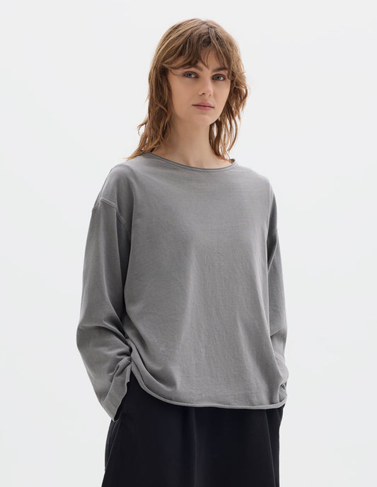 Cropped body image, front view of model wearing a wide neckline grey long sleeve T shirt. With dropped, wide sleeves with a loose cuff, finished with a rolled, raw edged hem. Worn over a black dress.