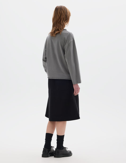 Full body image, back view of model wearing a  grey long sleeve T shirt. With dropped, wide sleeves with a loose cuff, finished with a rolled, raw edged hem. Worn over a black knee length dress and styled with black socks and black leather shoes.