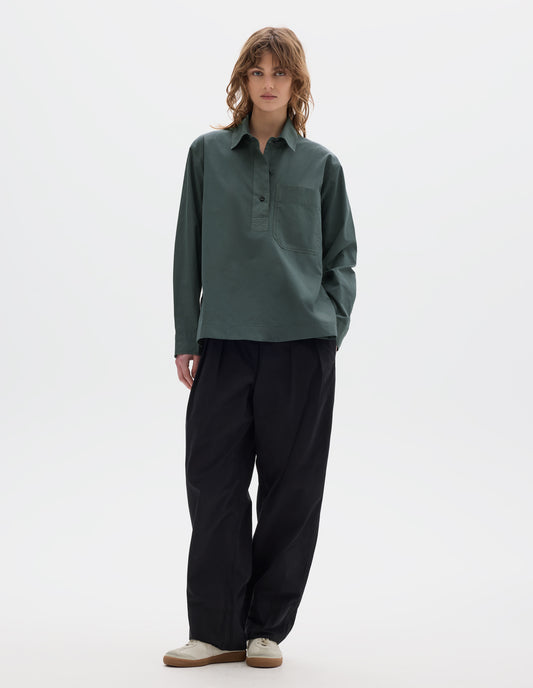 Full body image, front view of model wearing a greyish green cotton shirt with dark buttons, a half placket, chest patch pocket. Styled with black pleated trousers and off white suede trainers.
