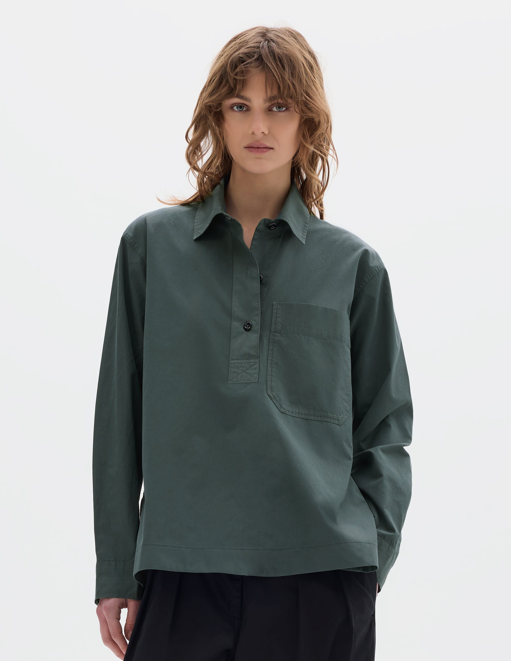 Cropped body image, front view of model wearing a greyish green cotton shirt with dark buttons, a half placket, chest patch pocket. Styled with black pleated trousers.