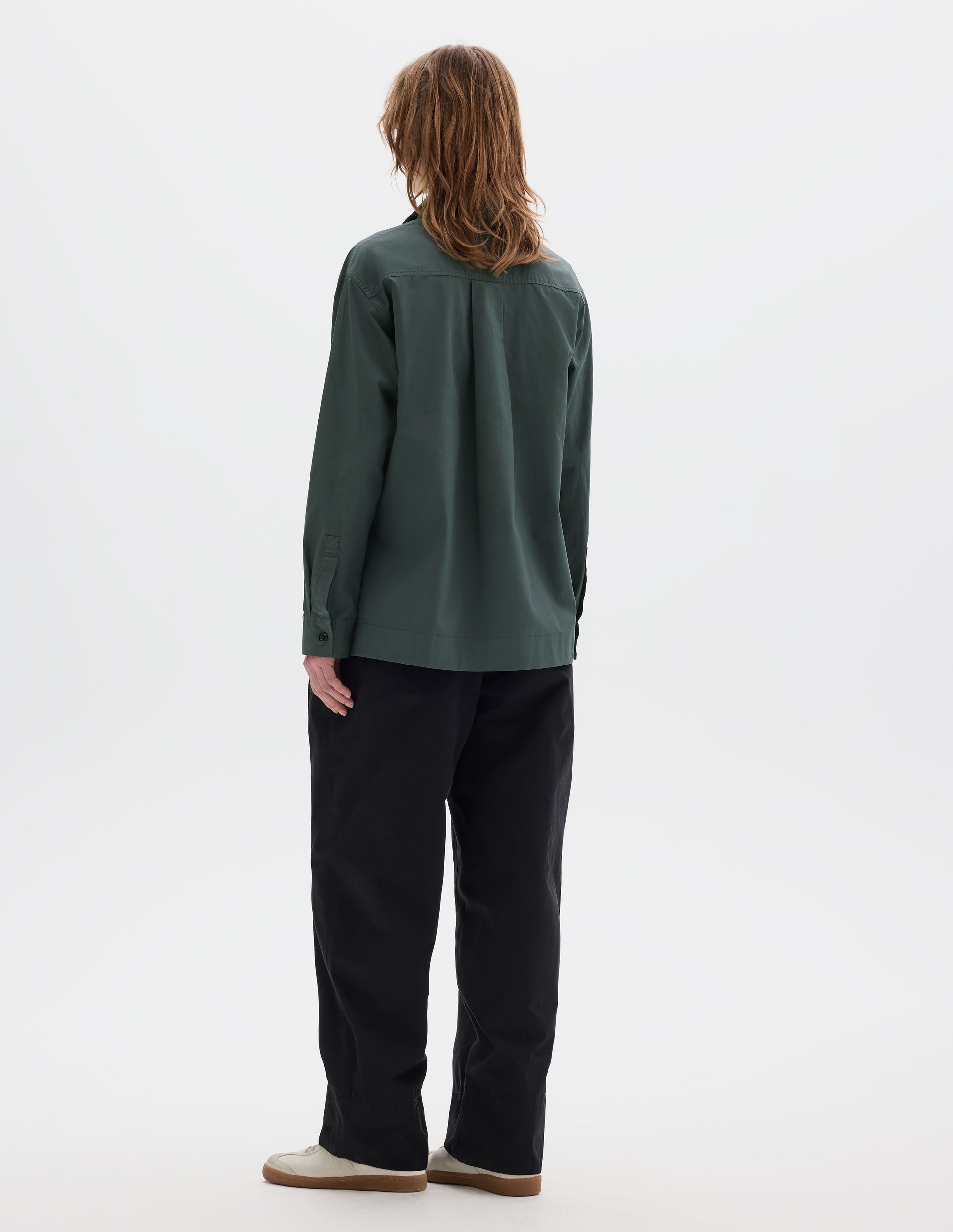 Full body image, back view of model wearing a greyish green cotton shirt with a box pleat on back yoke and a stepped hem. Styled with black pleated trousers and off white suede trainers.