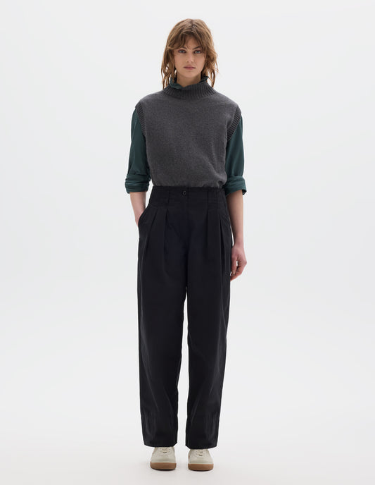 Full body image, front view of model wearing black cotton trousers, with front pleats, a brown button with belt loops and side pockets. Styled with blueish green shirt, grey slipover and white shoes.
