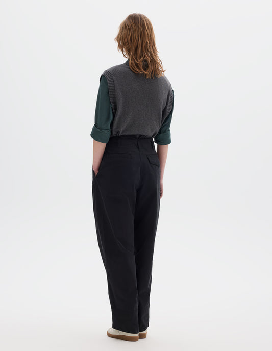 Full body image, back view of model wearing black cotton trousers, with front pleats, a brown button with belt loops and side pockets. Styled with blueish green shirt, grey slipover and white shoes.