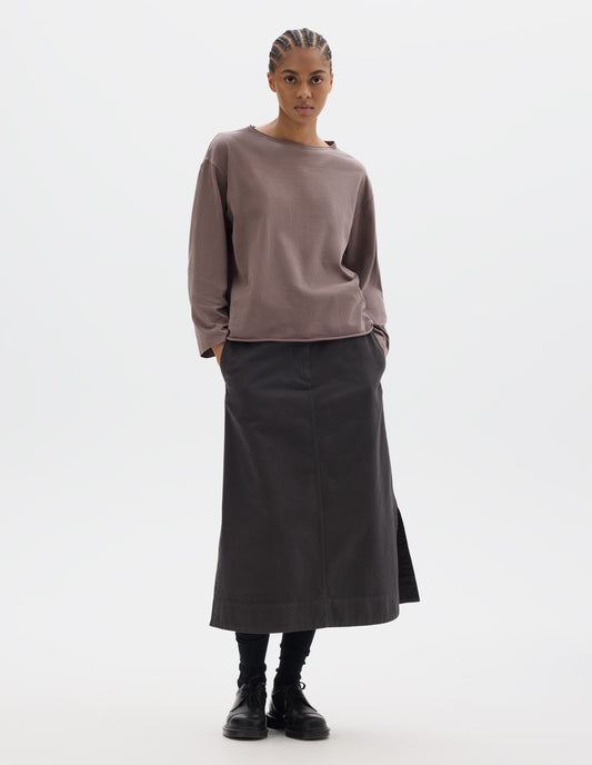 Full body image, front view of model wearing a wide neckline purpleish pink long sleeve T shirt. With dropped, wide sleeves with a loose cuff, finished with a rolled, raw edged hem. Styled with a longer dark brown skirt, black socks and black leather shoes.