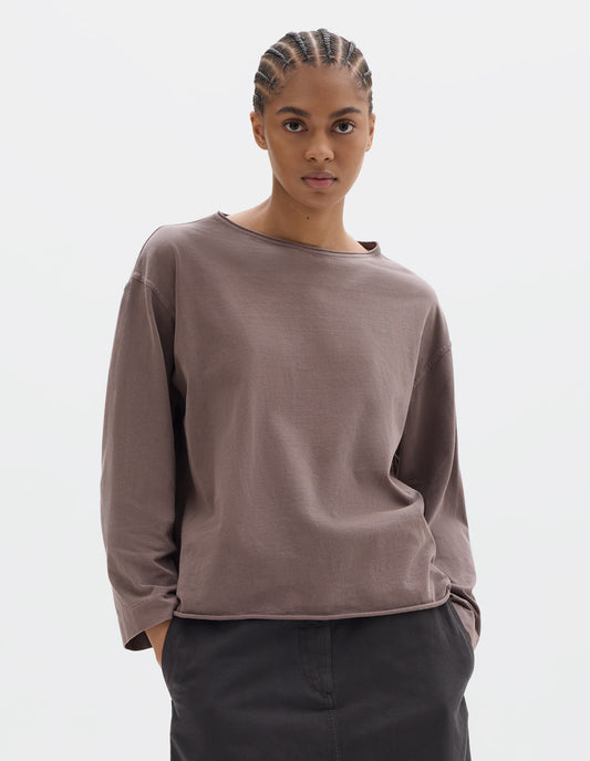 Cropped body image, front view of model wearing a wide neckline purpleish pink long sleeve T shirt. With dropped, wide sleeves with a loose cuff, finished with a rolled, raw edged hem. Styled with a dark brown skirt.