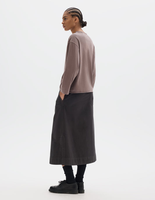 Full body image, back view of model wearing a wide neckline purpleish pink long sleeve T shirt. With dropped, wide sleeves with a loose cuff, finished with a rolled, raw edged hem. Styled with a longer dark brown skirt, black socks and black leather shoes.