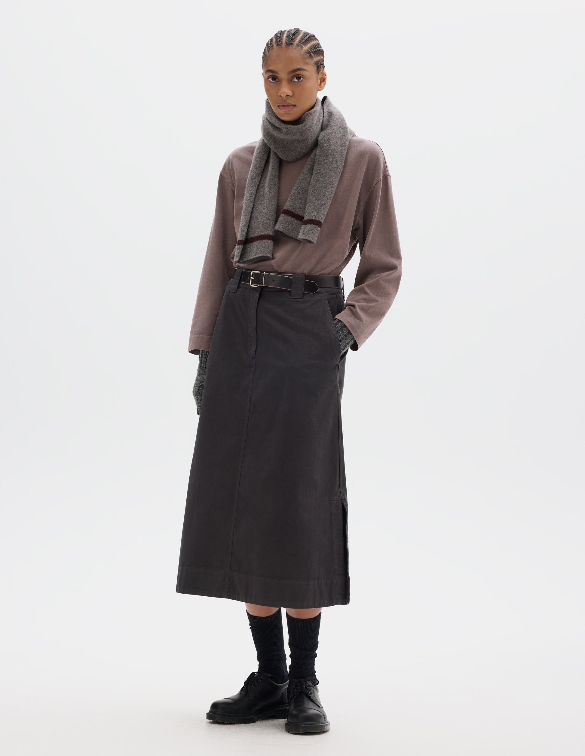 Full body image, front view of model wearing dark brown A line skirt with side splits, double hook and bar fastening waistband, belt loops and slant pockets. Styled with lilac long sleeve top tucked in, grey knitted scarf, black leather belt, black socks and black leather shoes.