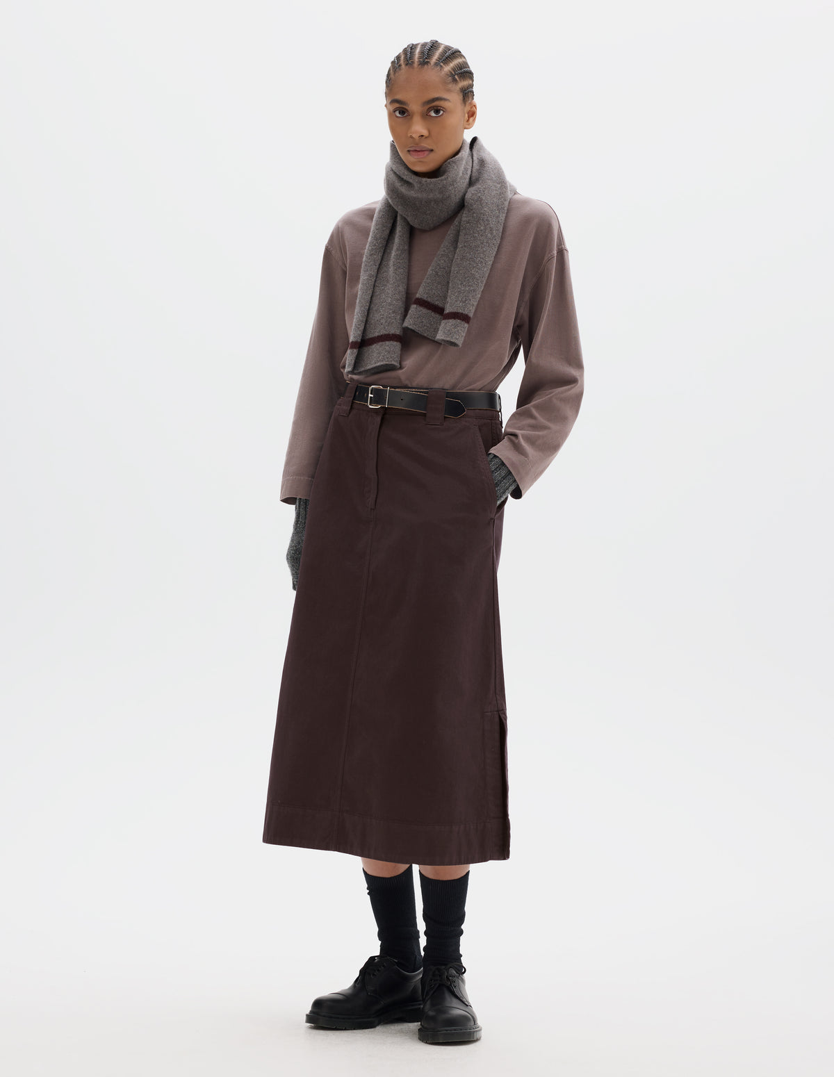 Full body image, front view of model wearing dark brown A line skirt with side splits, double hook and bar fastening waistband, belt loops and slant pockets. Styled with lilac long sleeve top tucked in, grey knitted scarf, black leather belt, black socks and black leather shoes.